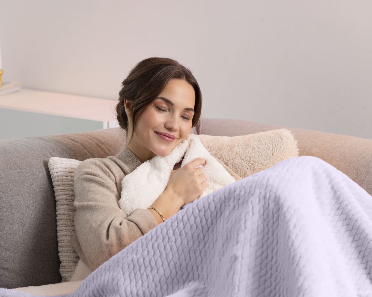 Frau kuschelt sich entspannt auf dem Sofa unter eine lilafarbene medisana Heizdecke.